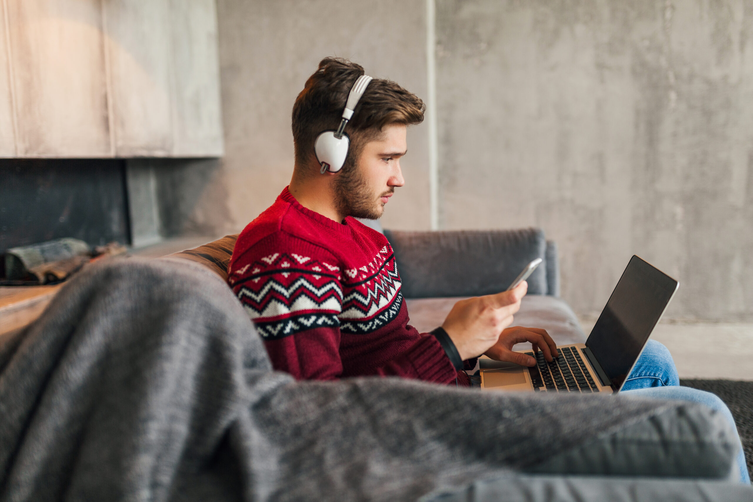 young attractive man on sofa at home in winter with smartphone in headphones, listening to music, wearing red knitted sweater, working on laptop, freelancer